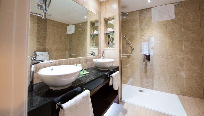 Brightly lit bathroom with two bowl sinks, a large mirror, a glass walled shower & light brown marbled tiles