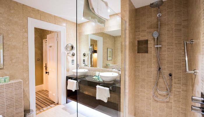 Brightly lit bathroom with two bowl sinks, a large mirror, a glass walled shower & light brown marbled tiles