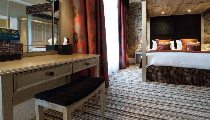 Side table with mirror, small chair in a room with large orange curtains & four post double bed