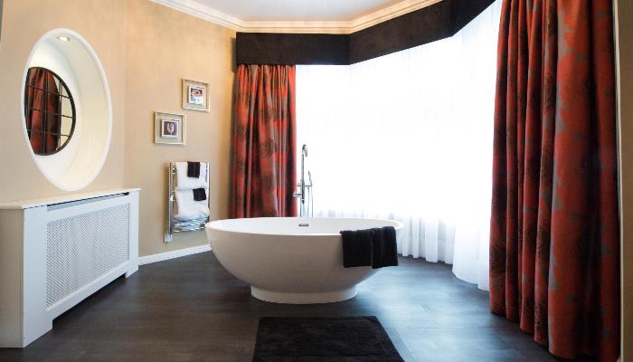 Freestanding bath by a large, wall length window with orange curtains & a port hole window looking into the adjoining bedroom area