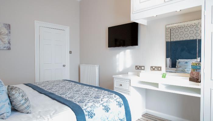 Bright room with white double bed decorated with a flowery blue blanket over the top with a mounted mirror & television on the opposite wall