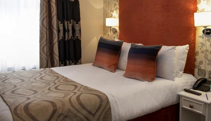 Double bed with white duvet, ligh brown patterned blanket with orange pillows & headboard flanked by wall lights.